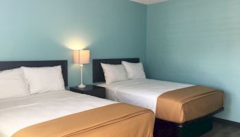 The image shows a hotel room with two double beds, white linens, brown blankets, a bedside table with a lamp, and light blue walls.