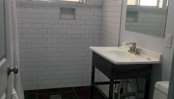 A small bathroom featuring a window, white tiles, a sink with a mirror above it, and a toilet.