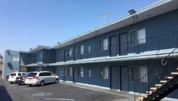 The image shows a two-story motel with blue walls. Several white and silver cars are parked nearby, and there are stairs on the right side.