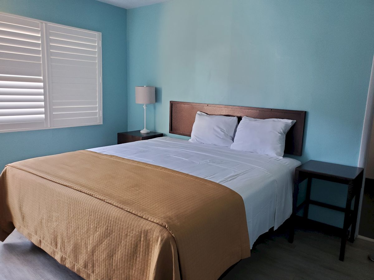 The image shows a neatly made bed with white linens and a tan blanket, flanked by two nightstands, in a room with light blue walls and a window with shutters.