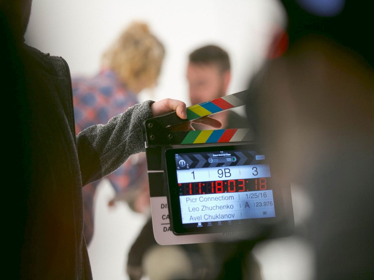 A film set scene with a hand holding a clapperboard in the foreground. Two blurred figures can be seen in the background.