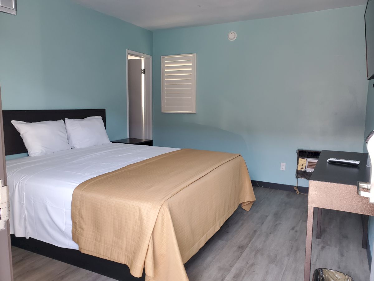 A neatly made bed with white sheets and a beige blanket in a minimalist room, featuring light blue walls and basic furniture, including a desk and chair.