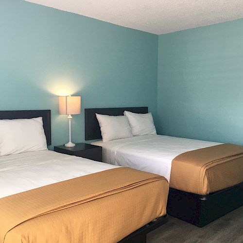 A clean, minimalist bedroom with two double beds, white bedding, and mustard-colored covers. A lit lamp is on a nightstand between the beds.