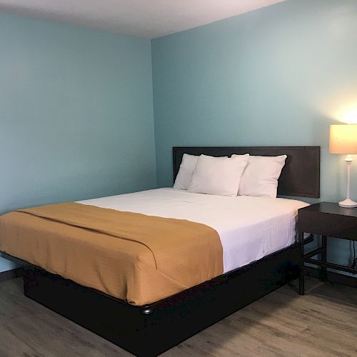 The image shows a neatly made bed with white pillows and an orange bedspread, next to a nightstand with a lit lamp in a room with light blue walls.