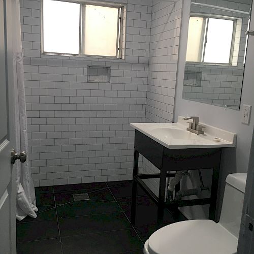 The image shows a bathroom with a white toilet, a sink with a mirror above, a black floor, and white tiled walls. There’s a window above the shower area.