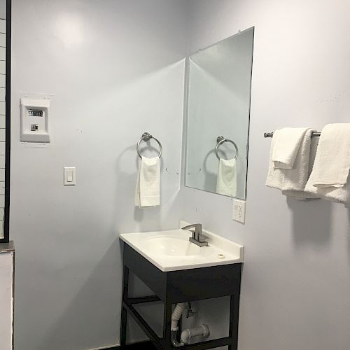 A clean bathroom with a sink, large mirror, hand towels, a trash can, and a towel ring on the wall. The decor is simple and neat.