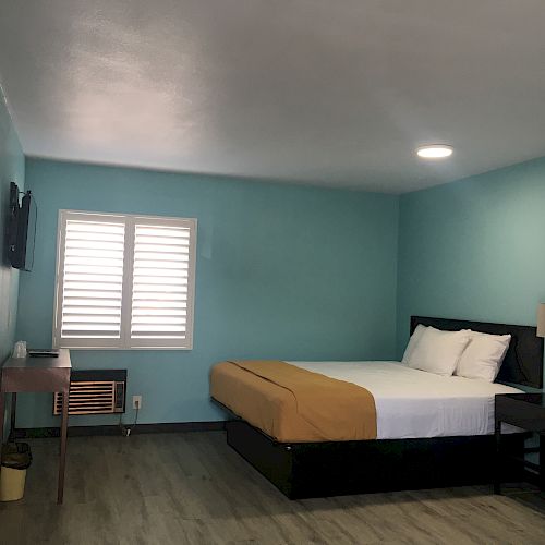 A neatly arranged room with a bed, pillows, nightstand, lamp, TV on the wall, air conditioning unit, and a window with shutters.