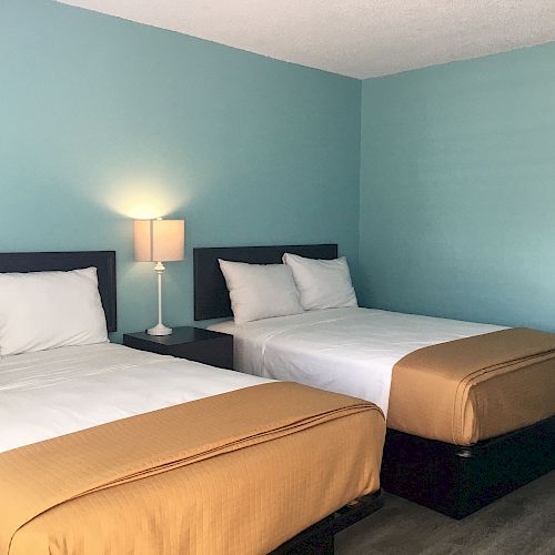 The image shows a hotel room with two neatly made double beds, a nightstand with a lamp between them, and light blue walls.