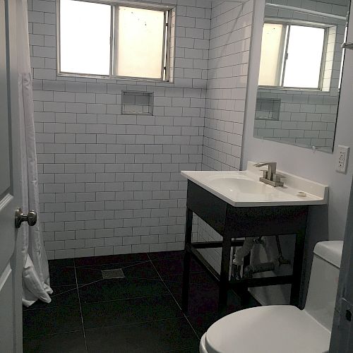 A bathroom with a white tiled shower, vanity with sink and mirror, toilet, window above, and dark floor tiles is shown in the image.