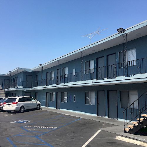 A two-story blue motel-like building with multiple doors and windows, a few parked cars, and a visible handicap parking space in the foreground.