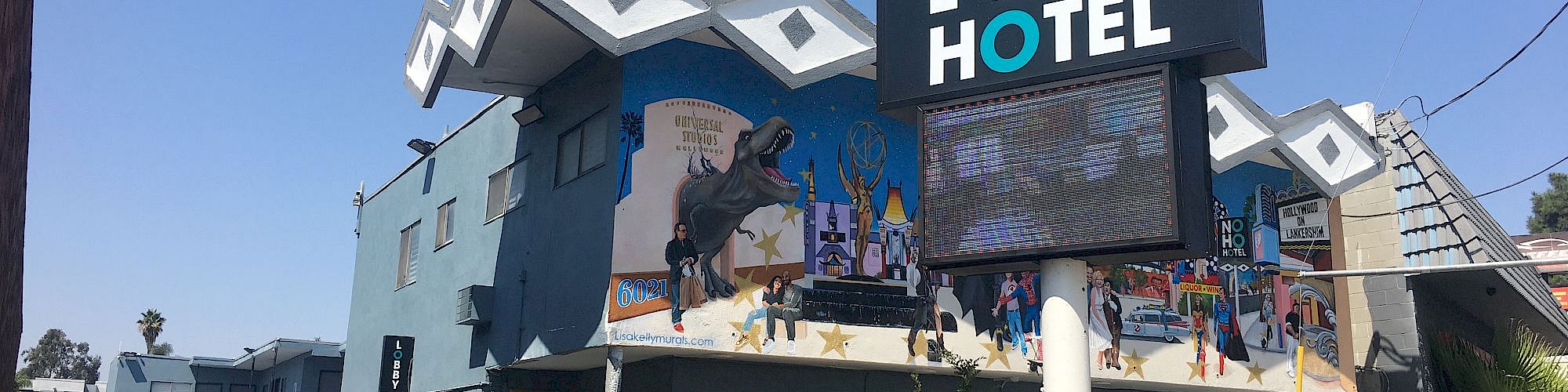 The image shows a blue NoHo Hotel building with a sign at the forefront. The building features a mural and a unique roof design.